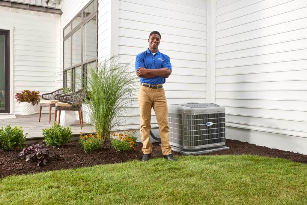 HVAC technician standing next to an air conditioning unit in front of a white house, smiling.