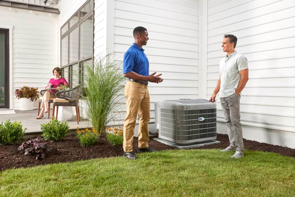 Two men discuss near AC unit outside house; woman relaxes on porch.