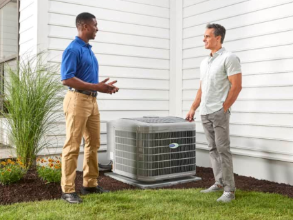 Two men near a gray AC unit outside a house. One in blue discusses, other in light shirt listens.