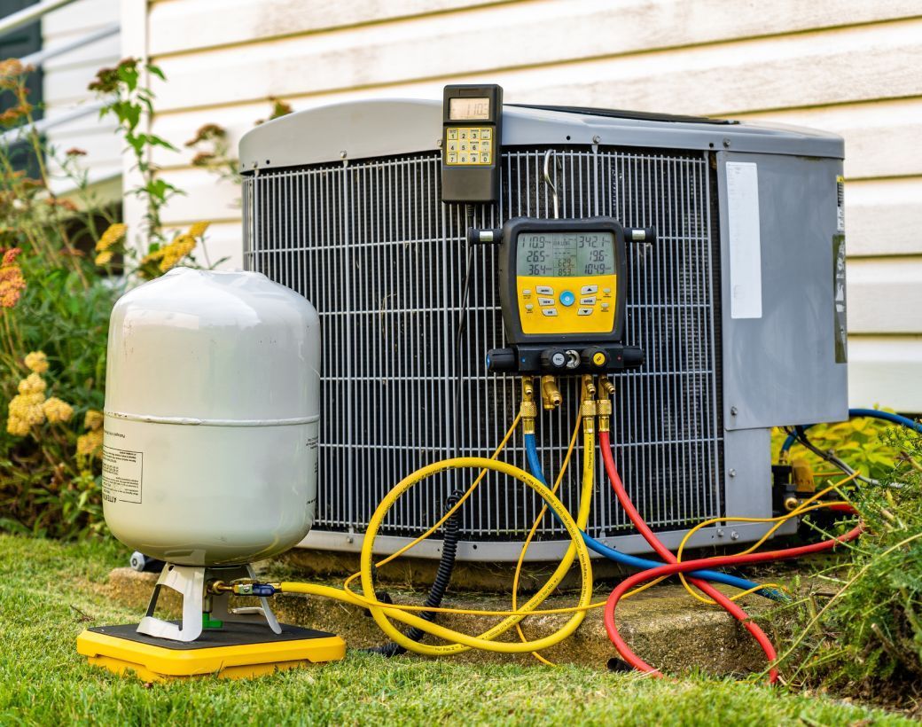 HVAC unit outdoors with gauges and yellow, blue, and red hoses connected. A white tank sits beside it.