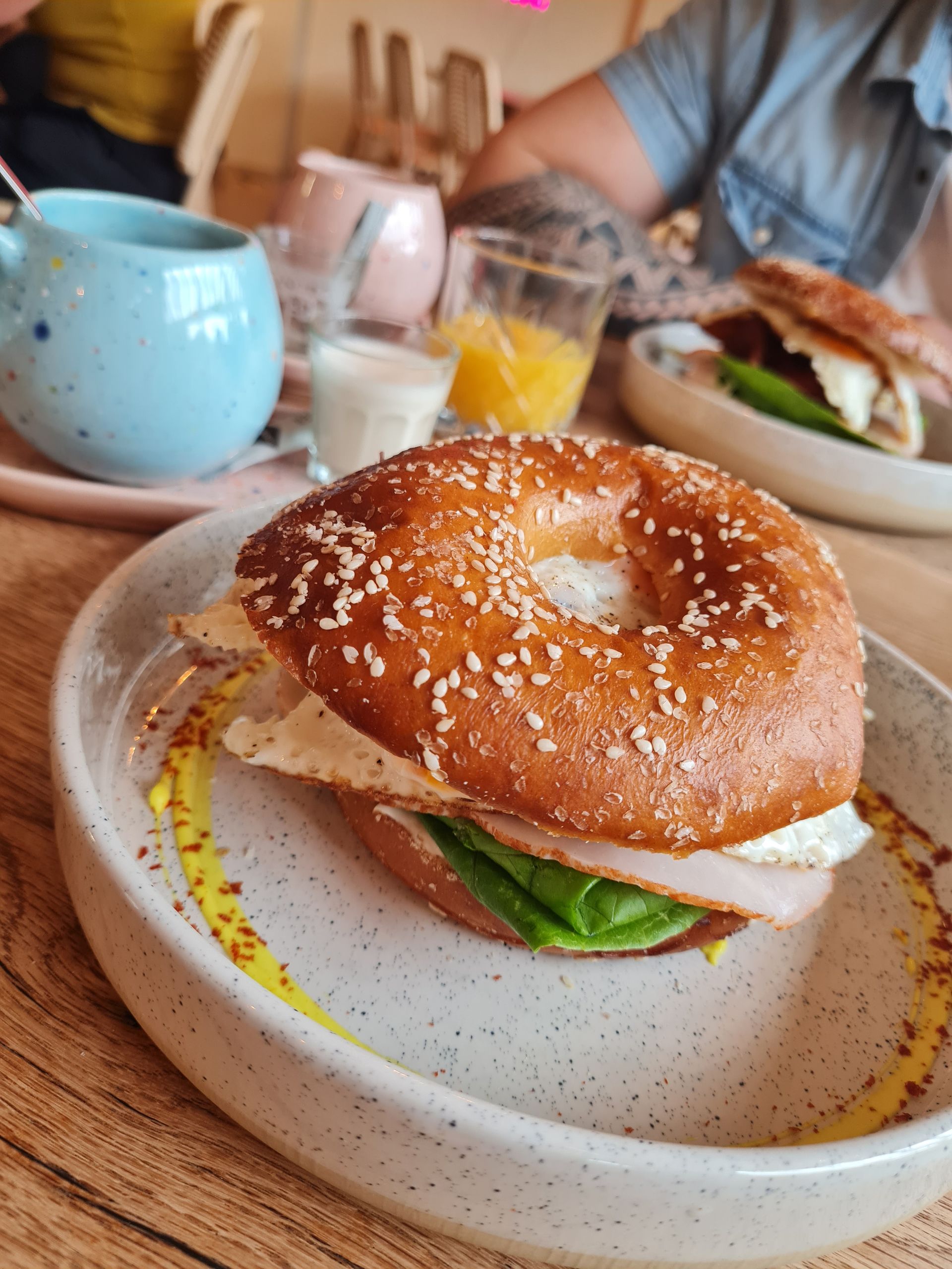 Bagel-Sandwich auf einem gesprenkelten Teller mit Ei, Schinken und Salat. Weitere Speisen und Getränke auf dem Tisch. Brunch mit Kunden.