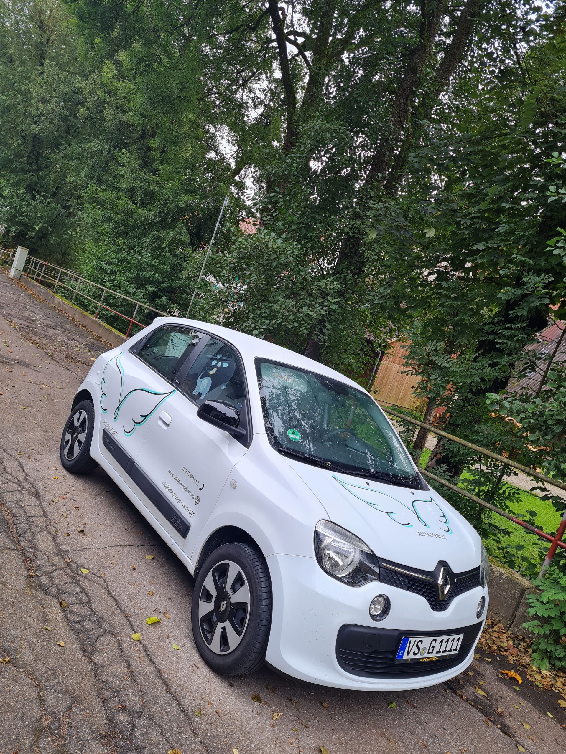 Ein weißer Renault Twingo parkt an einer abschüssigen Straße; im Hintergrund sind Bäume zu sehen. Das ist das Dienstauto.