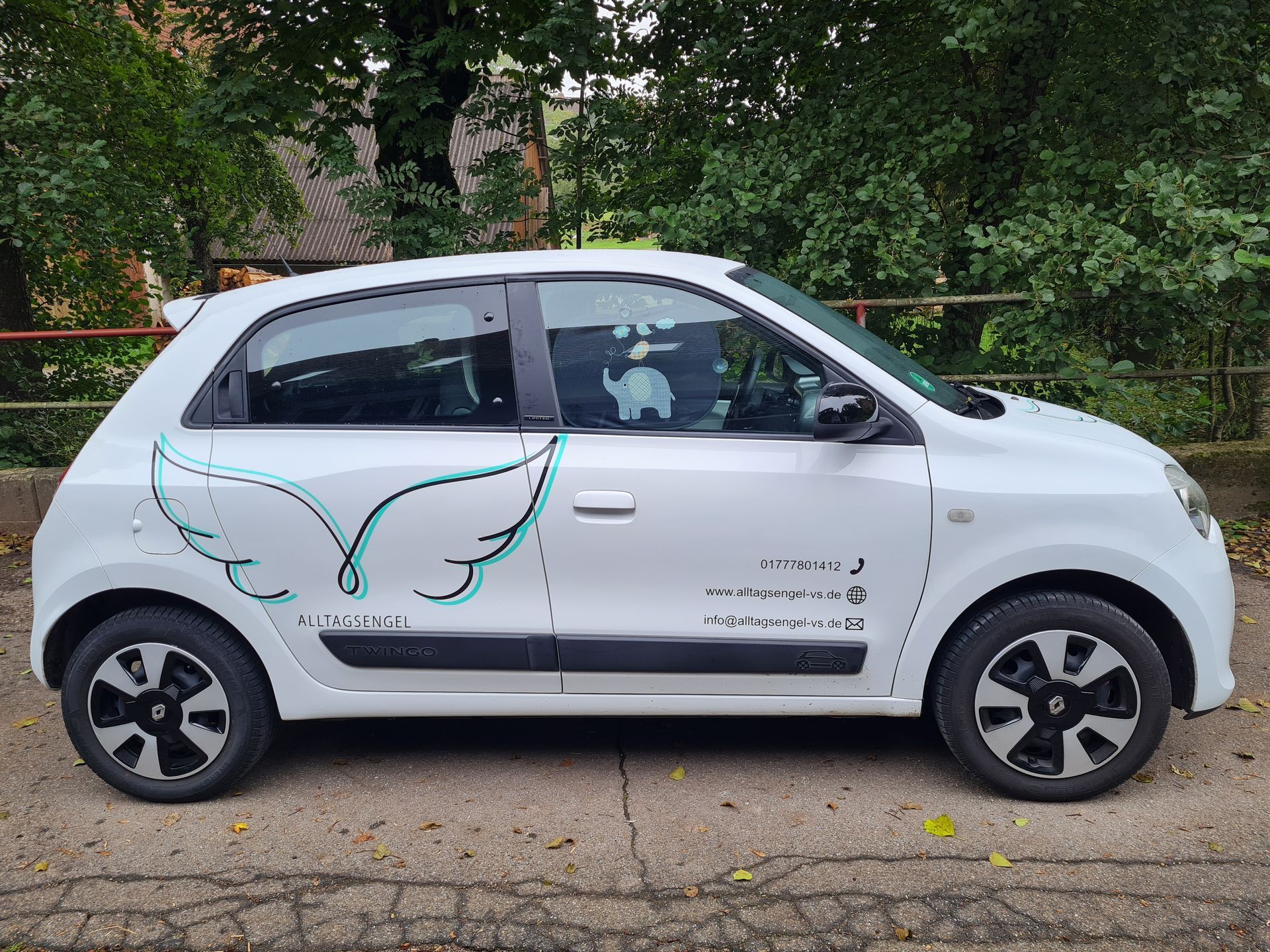 Ein weißer Renault Twingo mit türkisfarbenem Kotflügelaufkleber parkt auf einer Straße mit Bäumen im Hintergrund. Das ist das Dienstauto.