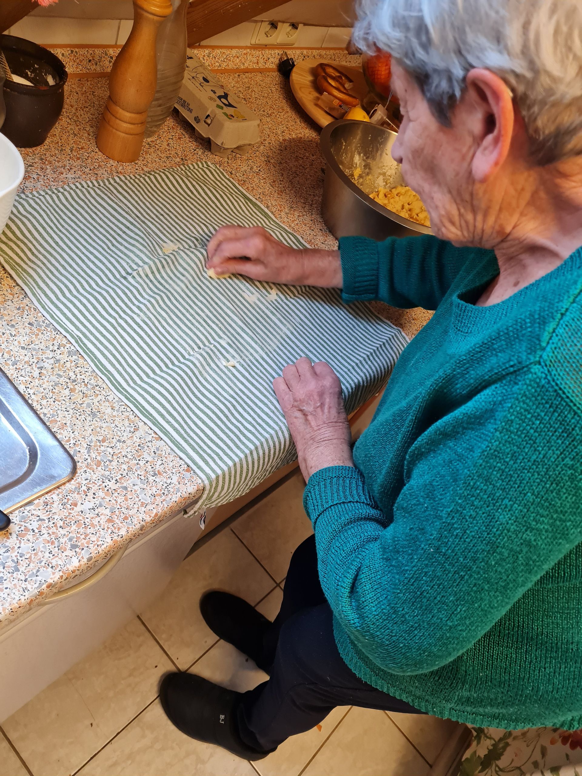 Person in grünem Pullover an einer Küchentheke, die Hände auf einem gestreiften Handtuch. Serviettenknödl entstehen.