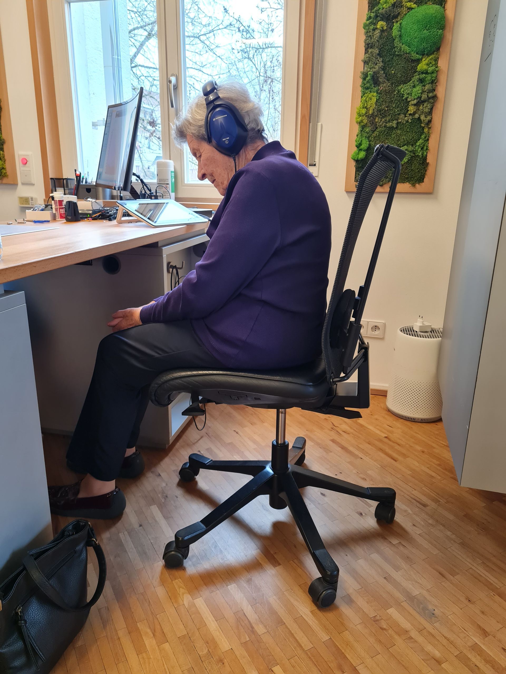 Frau in lila Pullover, mit Kopfhörern, sitzt an einem Schreibtisch mit Computer und Büromaterial. Hörtest Begleitung.