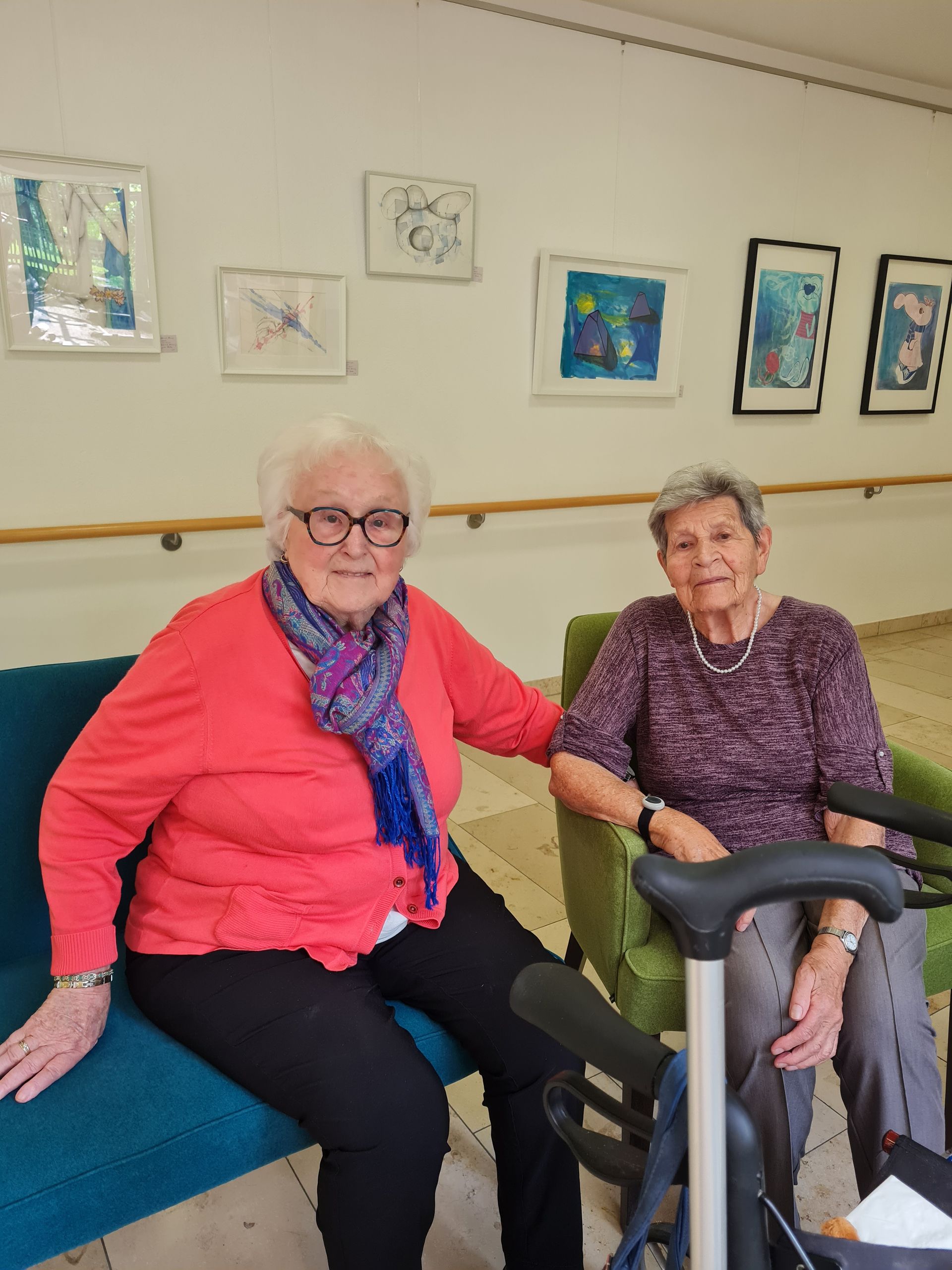 Zwei ältere Frauen sitzen neben einem Kunstwerk an der Wand. Eine ist in Rot, die andere in Lila gekleidet.