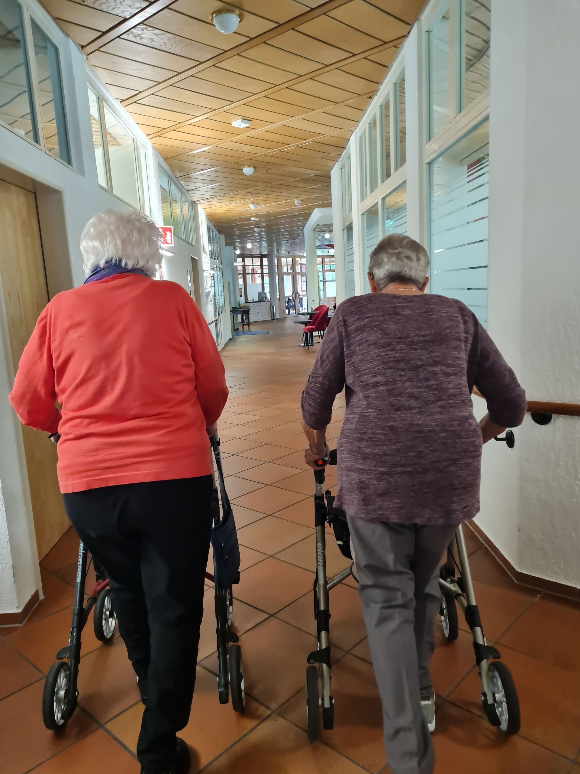 Zwei Personen, die Gehhilfen benutzen, gehen in einem langen Flur von der Kamera weg. Freundin im Pflegeheim besuchen.