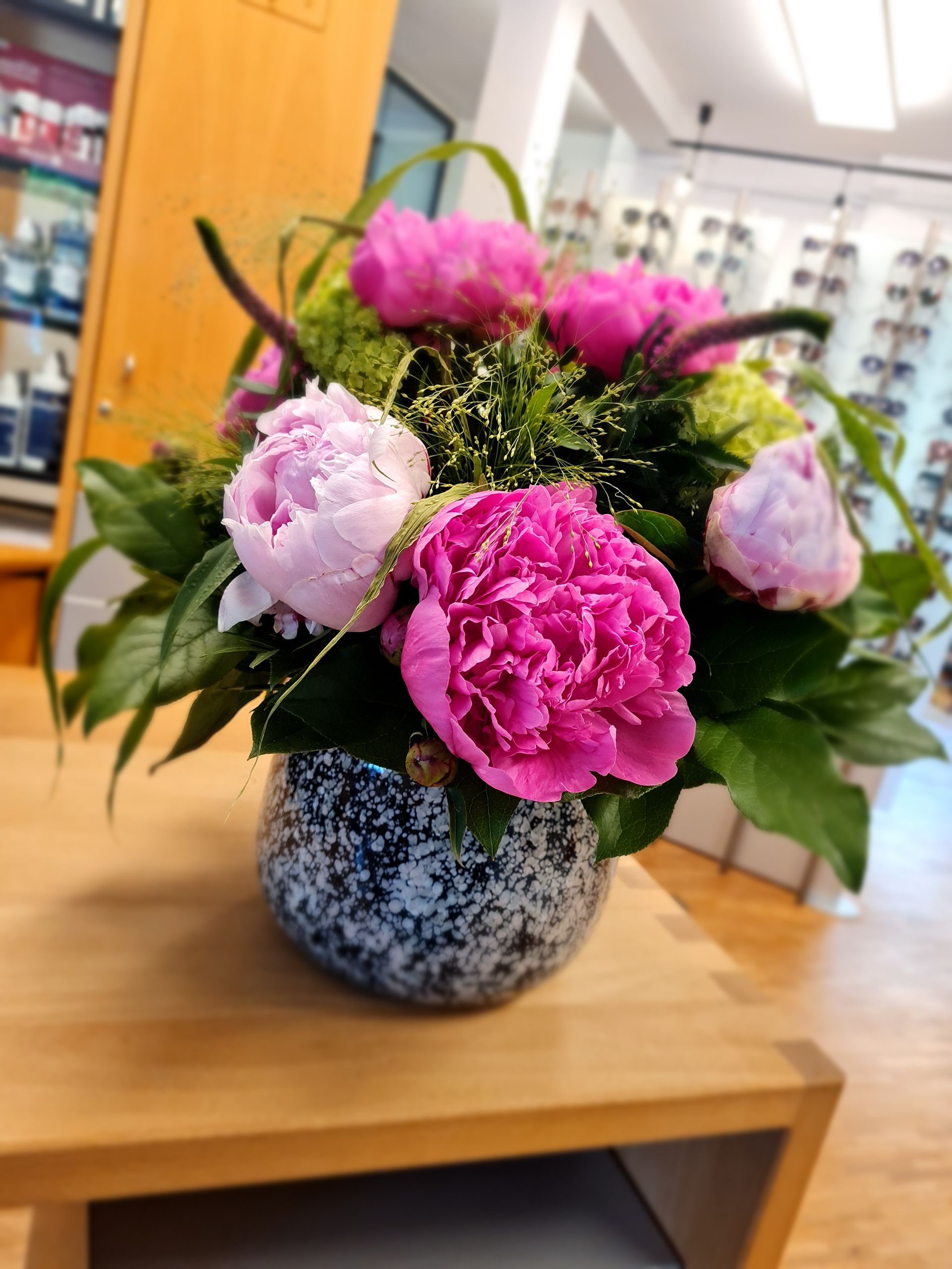 Ein Strauß rosa Pfingstrosen in einer gesprenkelten blauen Vase auf einem Holztisch, Ladenambiente.