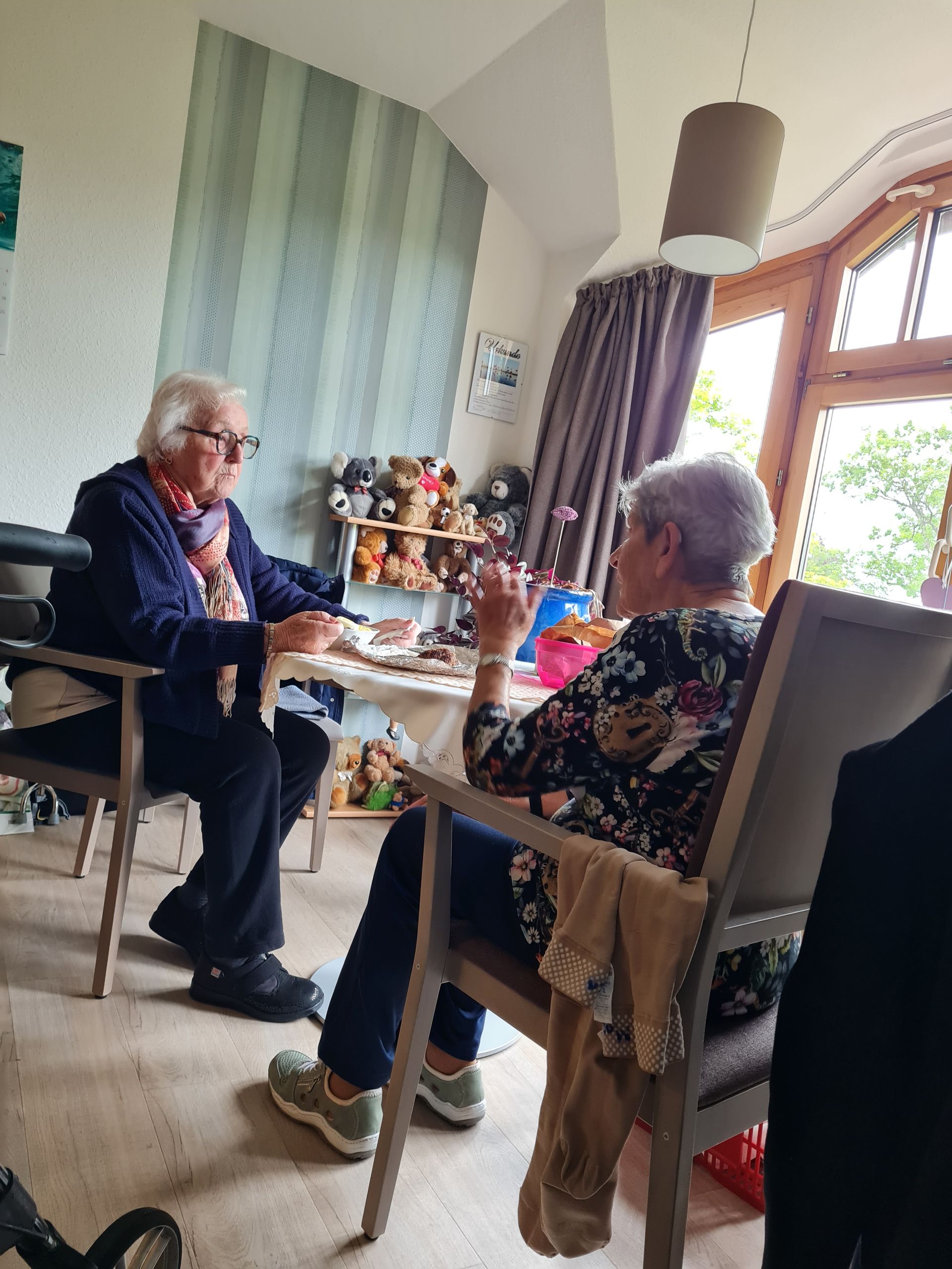 Zwei ältere Erwachsene sitzen an einem Tisch und unterhalten sich. Einer trägt eine Brille und einen blauen Pullover. Das Gespräch findet in einem Innenraum in der Nähe eines Fensters statt.