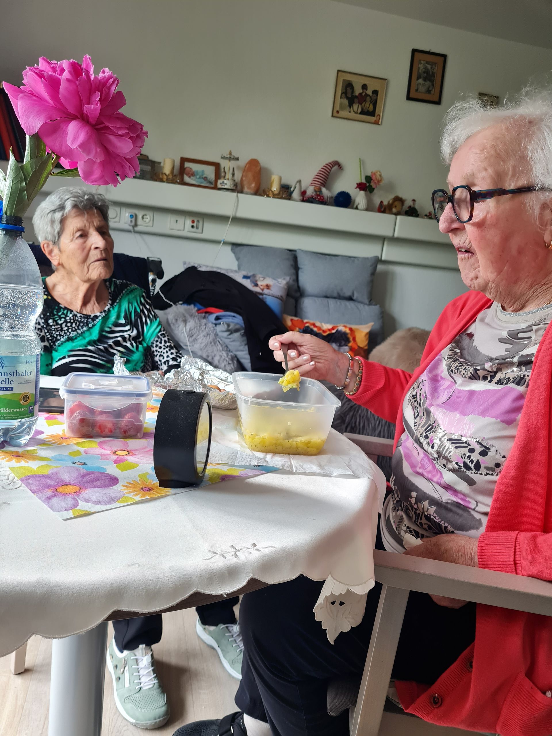 Zwei ältere Damen sitzen drinnen an einem Tisch. Die eine isst aus einer Schüssel, die andere schaut zu. Rosa Blumen und Speisen stehen auf dem Tisch. Besuch im Altenheim.