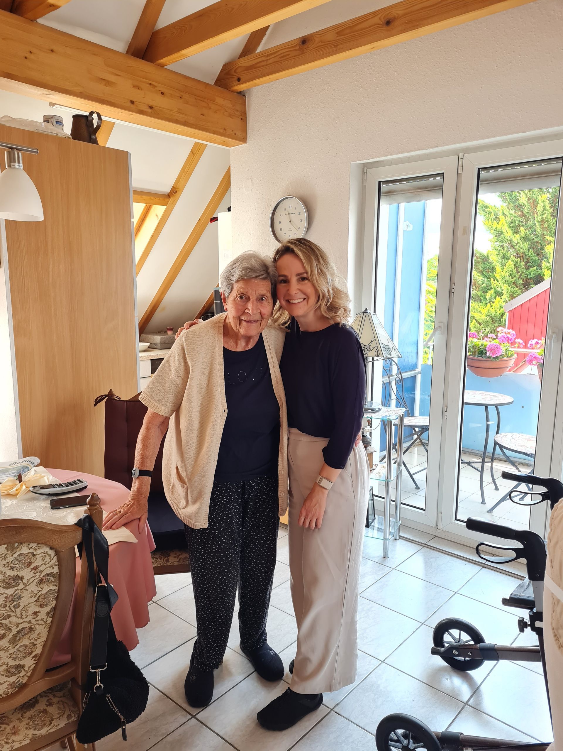 Frau und ältere Person lächeln drinnen, stehen an einer Balkontür. Herta und Tamara