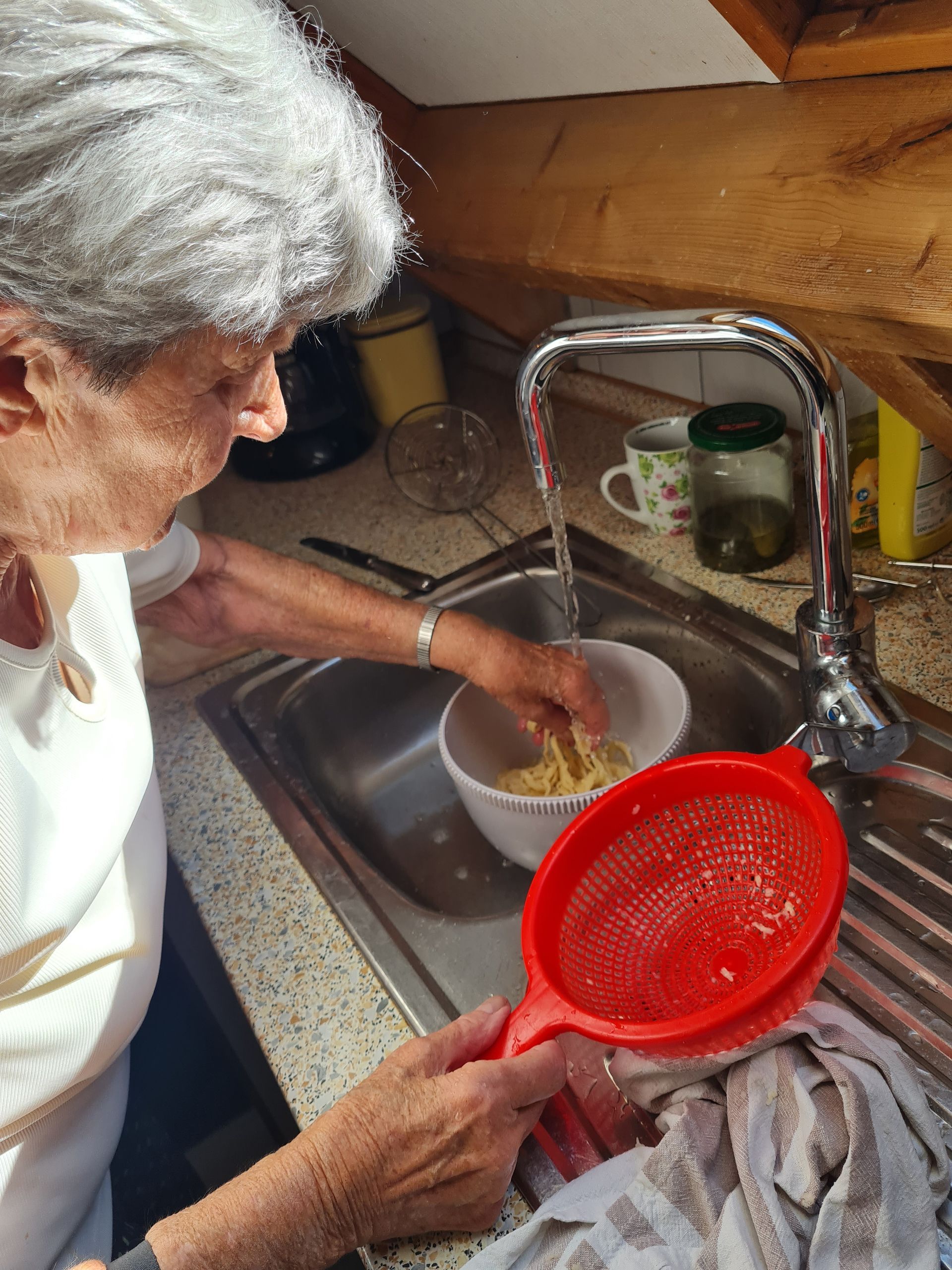 Eine Frau spült Lebensmittel in einer weißen Schüssel unter einem Wasserhahn ab und hält dabei ein rotes Sieb in der Küchenspüle. Gemeinsames Kochen und Abwaschen.