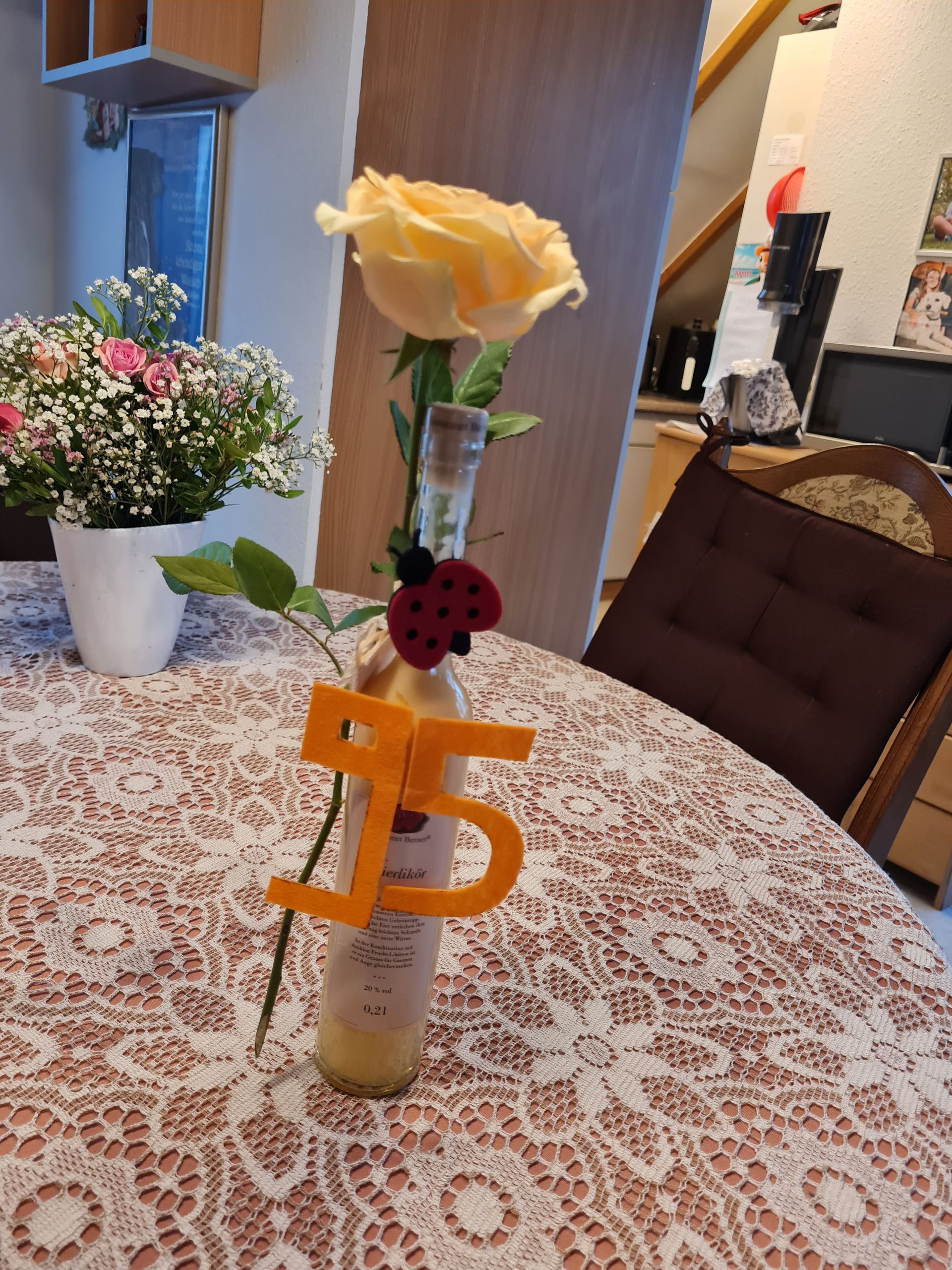 Gelbe Rose in einer Flasche mit der Aufschrift „95“ auf einer Spitzentischdecke; im Hintergrund ein Blumenarrangement und ein Stuhl. Geburtstagsgeschenk.