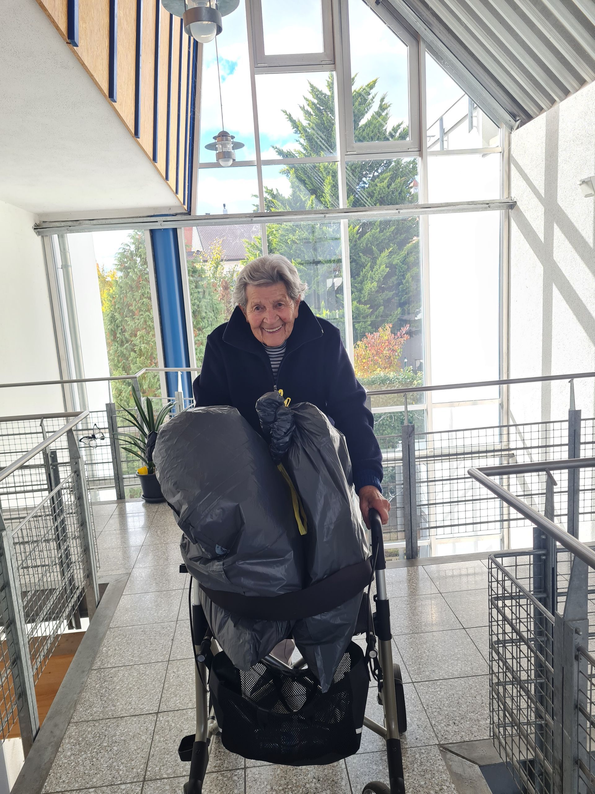 Eine ältere Person schiebt einen Rollator mit Taschen darin auf einem hellen, gefliesten Flur.