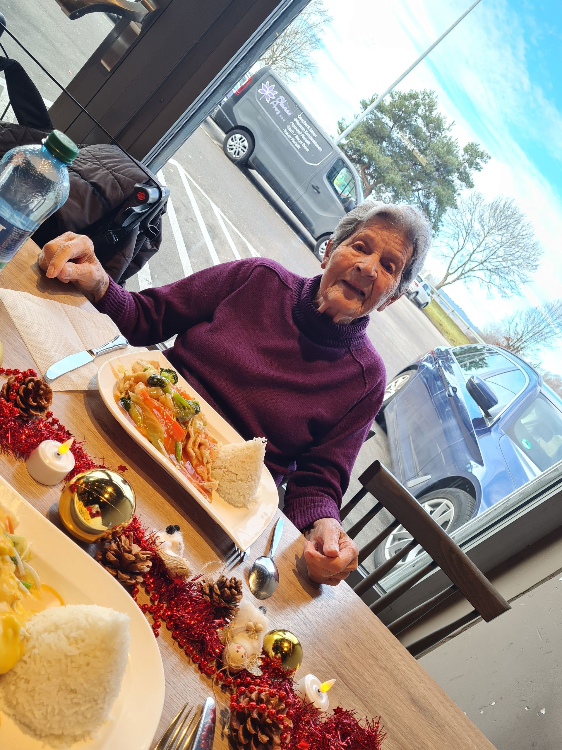 Eine ältere Person lächelt glücklich an einem mit Speisen und festlicher Dekoration gedeckten Tisch am Fenster.