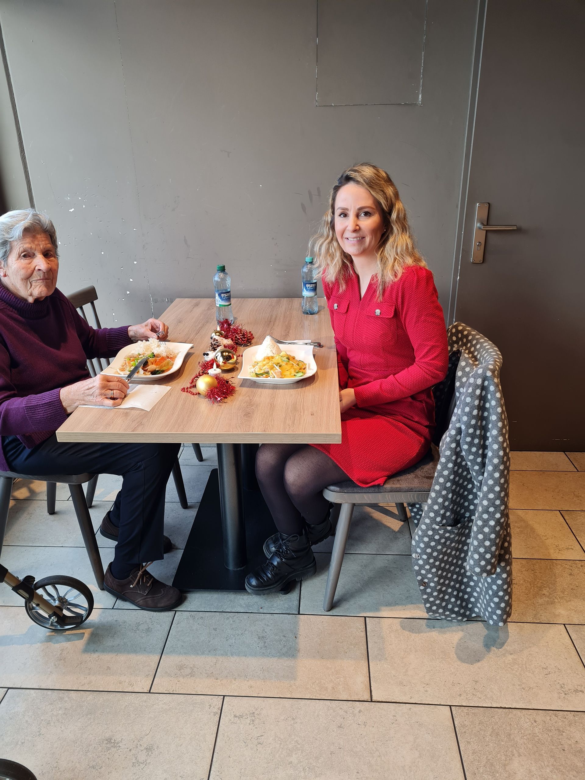 Zwei Frauen sitzen in einem Café an einem Tisch und essen. Die eine trägt ein rotes Kleid, die andere ein violettes Hemd. Speisen und Getränke stehen auf dem Tisch. Weihnachtsfeier mit 95 Jährigen Herta.