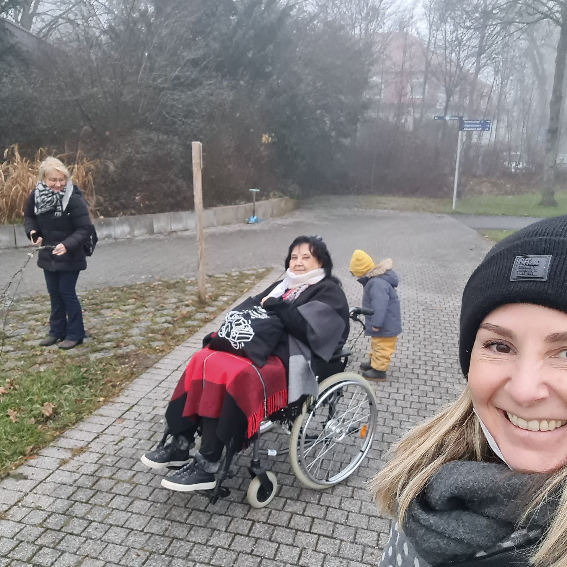 Frau im Rollstuhl auf dem Weg mit zwei anderen Personen, eine davon mit Kind, an einem nebligen Tag. Kurgarten in Bad Dürrheim.