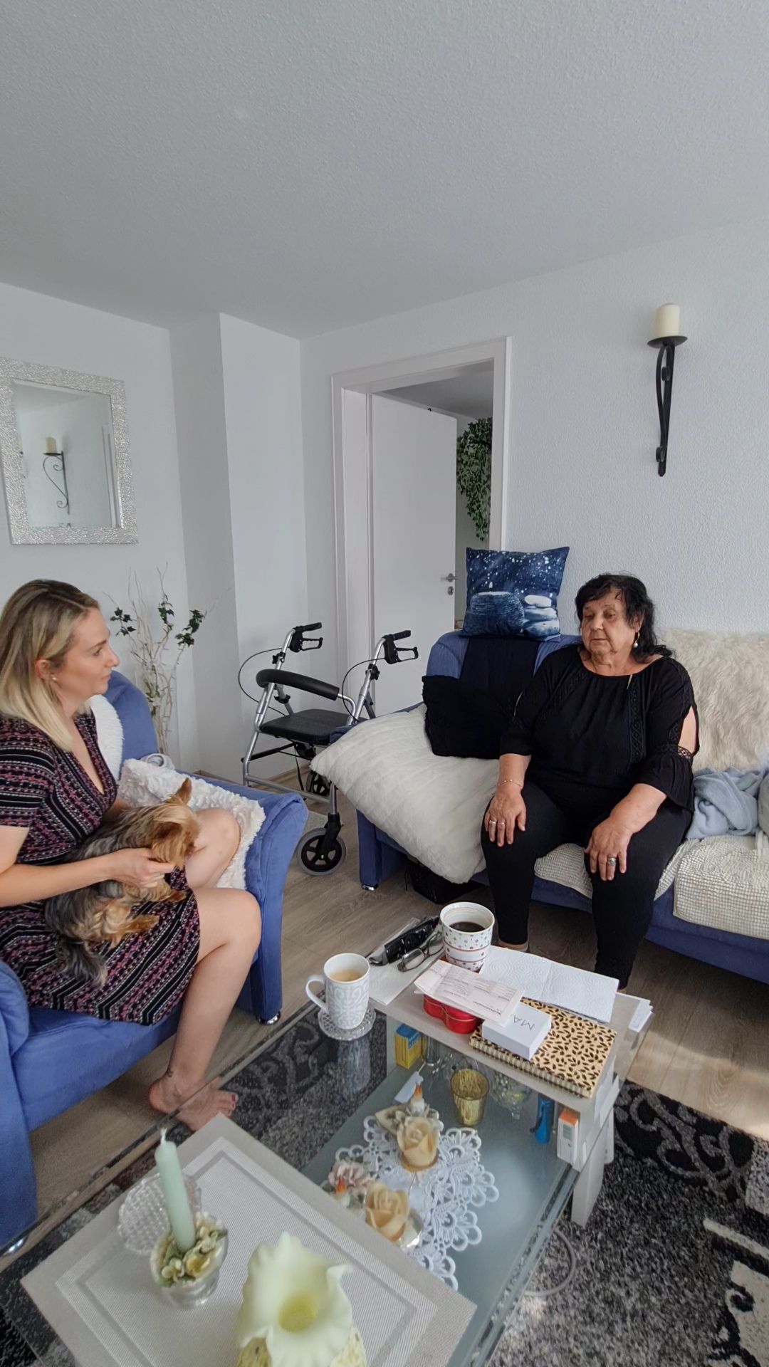 Eine Frau sitzt mit einem kleinen Hund auf einem blauen Sessel und unterhält sich mit einer anderen älteren Frau auf einem Sofa. Davor steht ein Couchtisch mit Snacks.