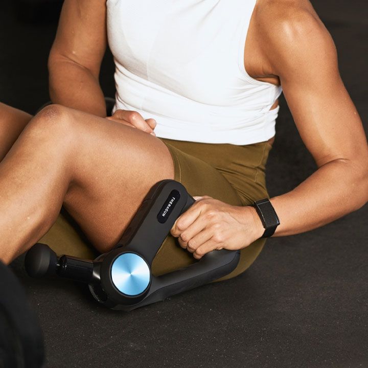 A man is sitting on the floor holding a massager that says theragun on it