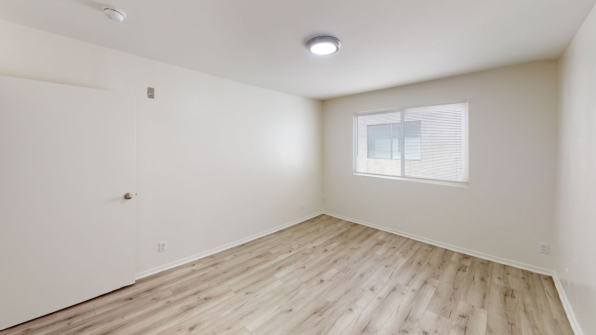 Empty room with light wood floor, white walls, small window, and closed door.