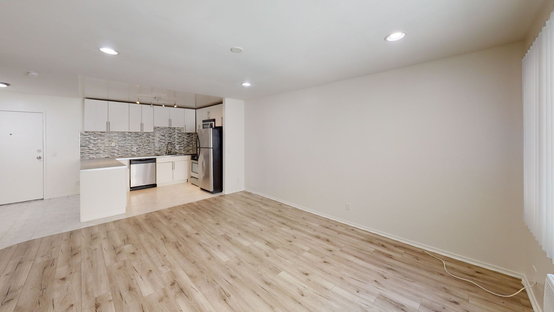 Empty apartment interior with kitchen in background; light wood floors, white walls, stainless steel appliances.