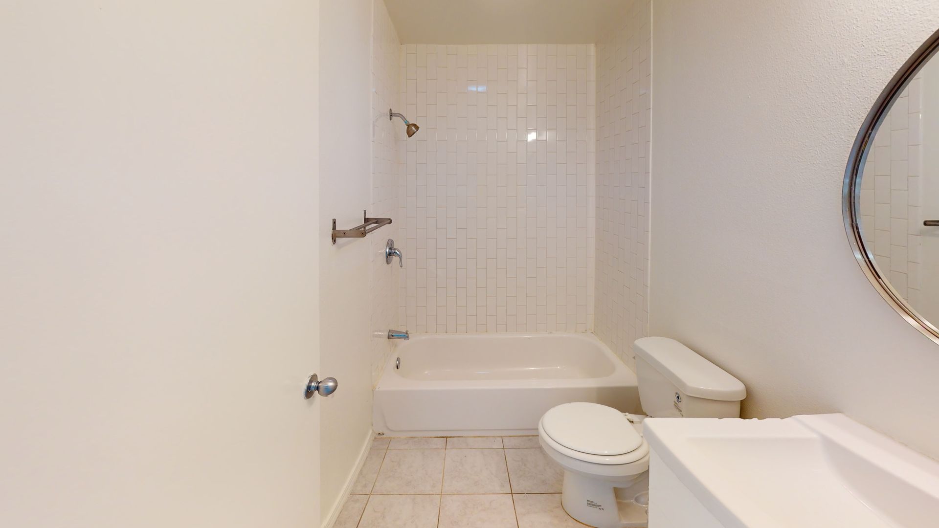 Bathroom with white tiled shower/tub, toilet, sink, and round mirror.