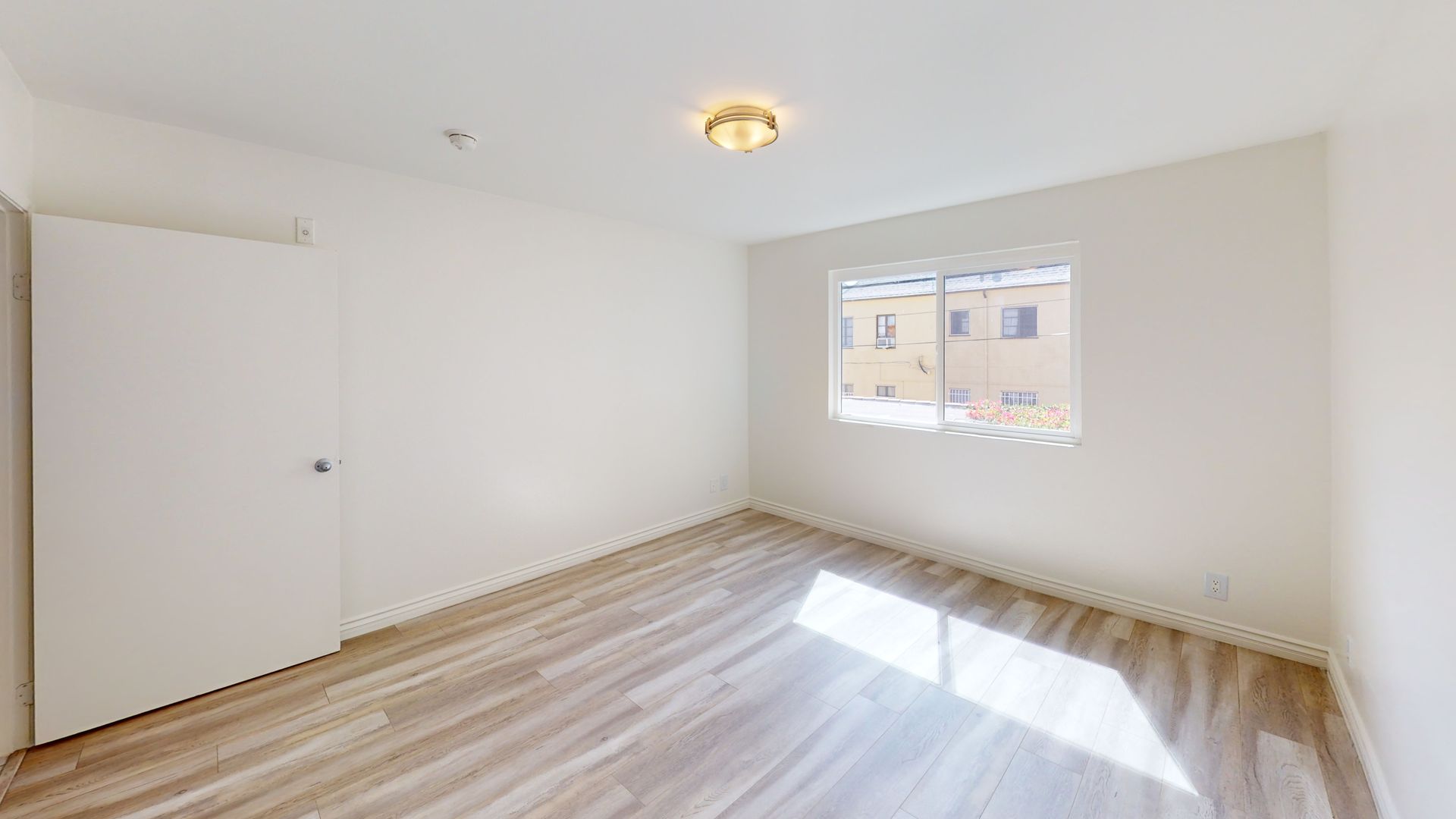 Empty, bright bedroom with light wood flooring, a closed door, and a small window.