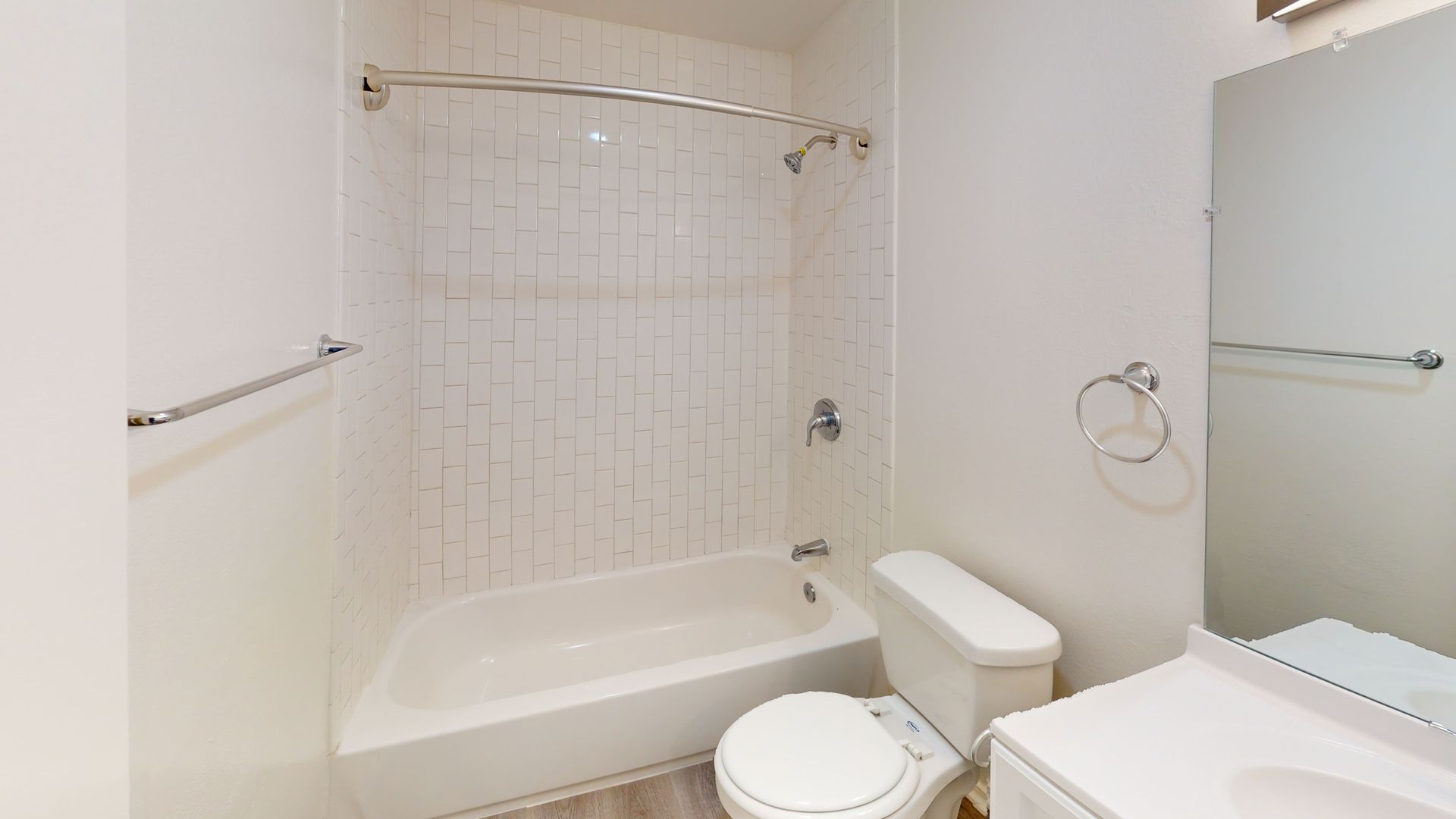 White bathroom with bathtub, toilet, sink, and mirror.  Tiled shower wall, chrome fixtures, light wood floor.