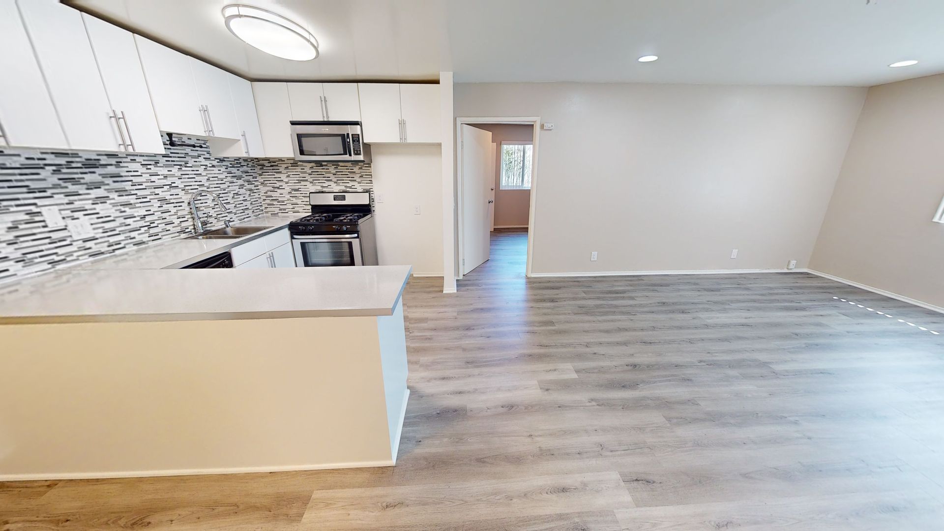 Modern kitchen and living area, white cabinets, stone backsplash, gray flooring. Doorway visible in the background.