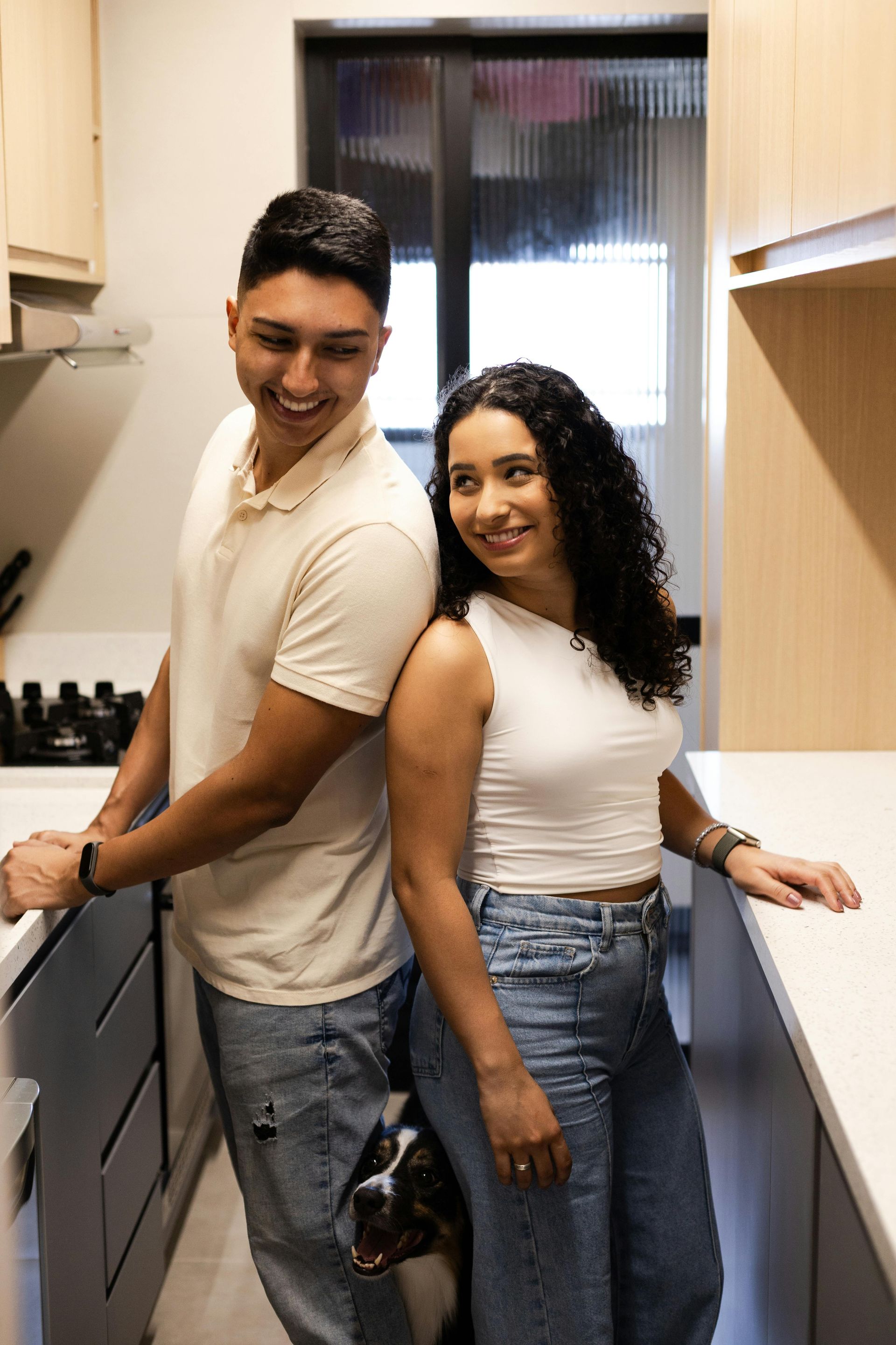Couple standing in a kitchen, smiling. Woman in white top, jeans. Man in polo shirt, jeans. Dog at their feet.