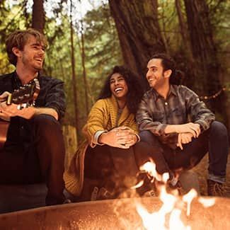 Three people smiling around a campfire in a forest; one plays a guitar.