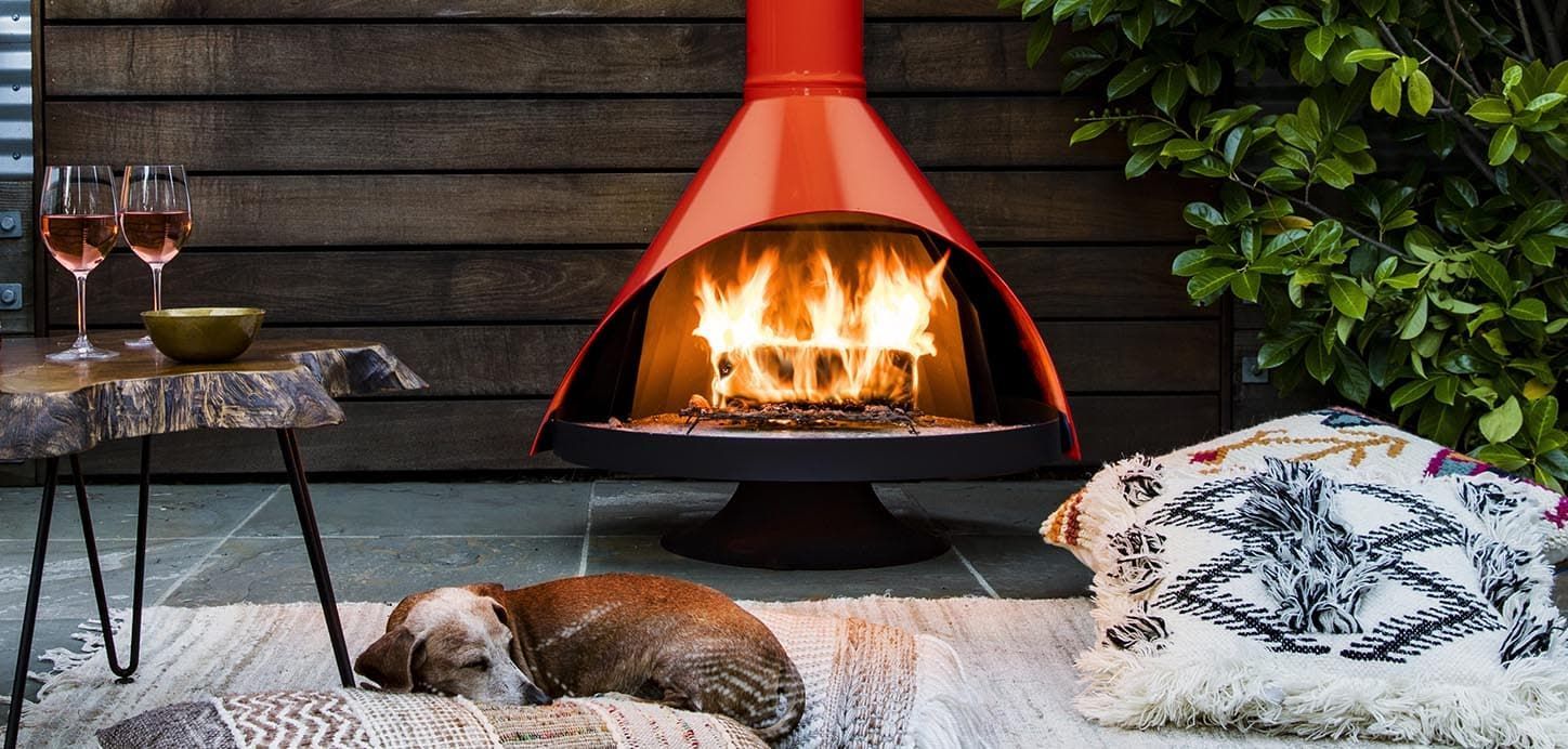 A cozy outdoor scene with a dog sleeping by a fireplace with flames. Two wine glasses and a pillow are nearby.