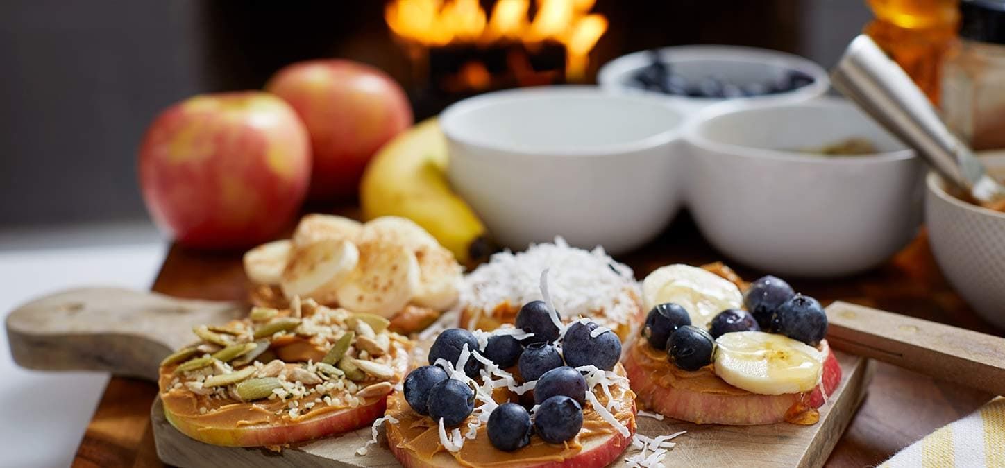 A wooden cutting board topped with apples , bananas , blueberries and peanut butter.