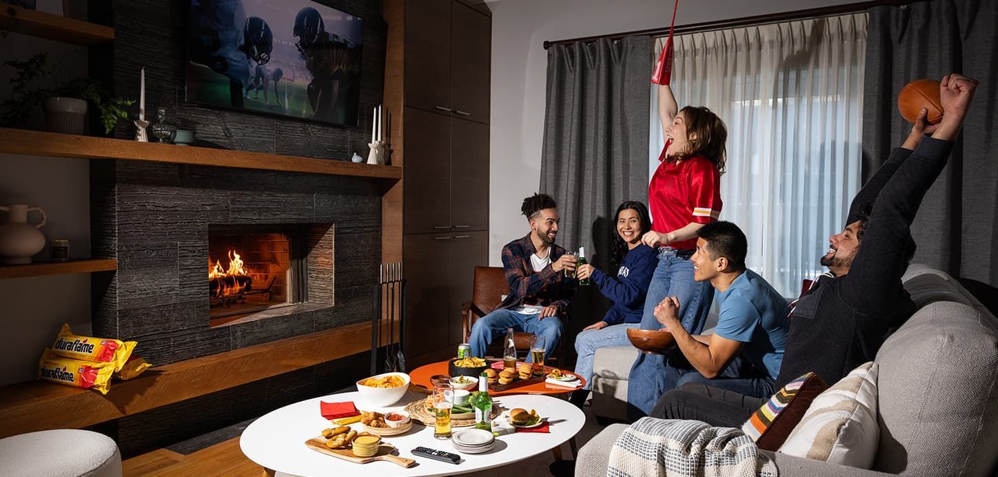 People watch TV in a living room, some cheer. Food and drinks are on the coffee table. Fireplace in background.