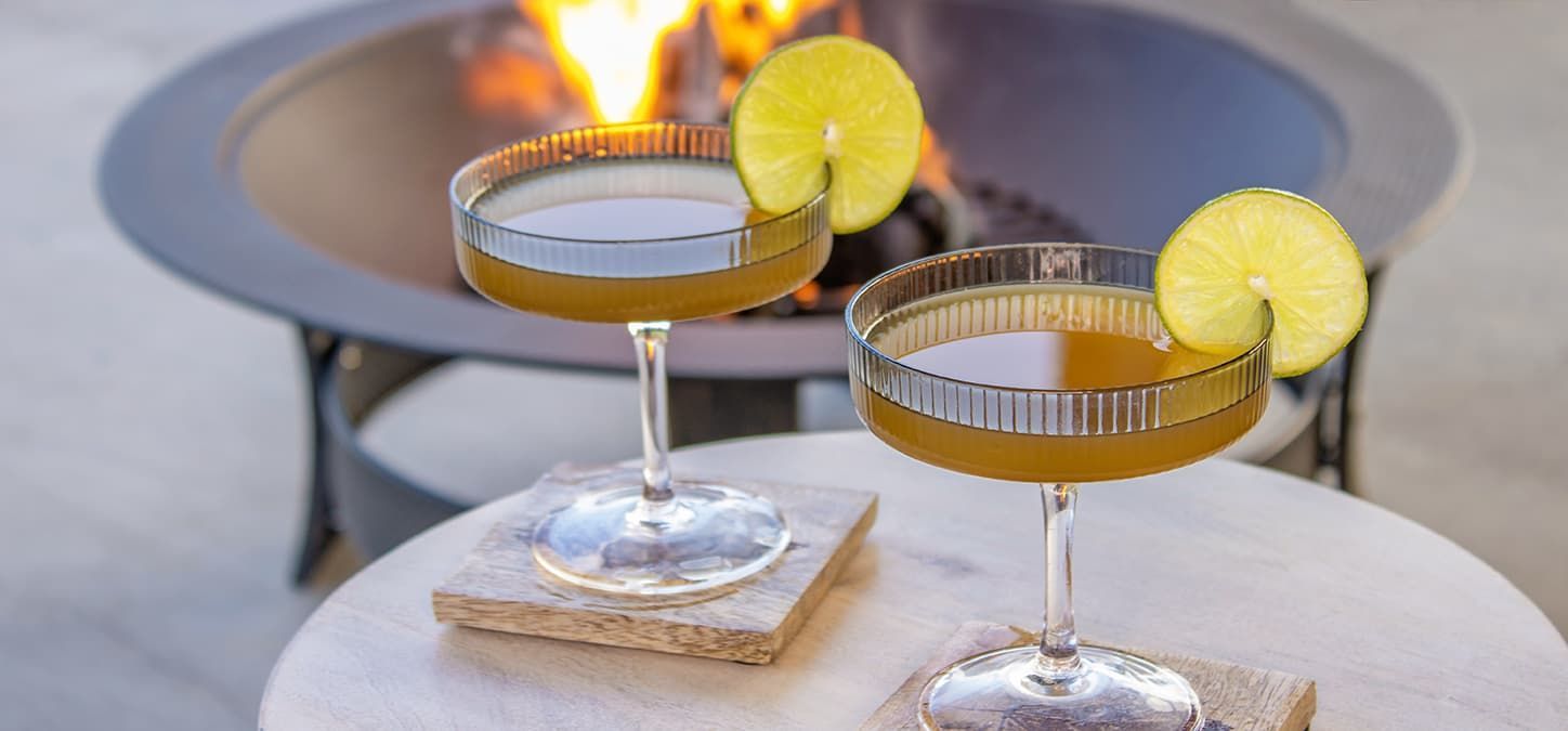 Two cocktails are sitting on a table in front of a fire pit.