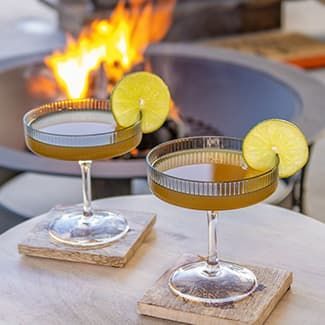 Two martini glasses with lemon slices on them are sitting on a table in front of a fire pit.