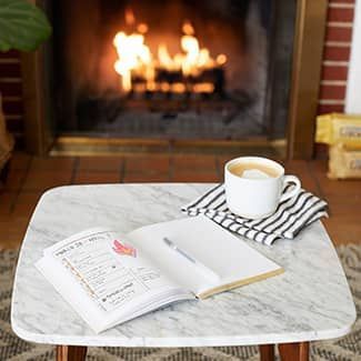 Marble table with notebook, coffee cup, and fireplace in the background.