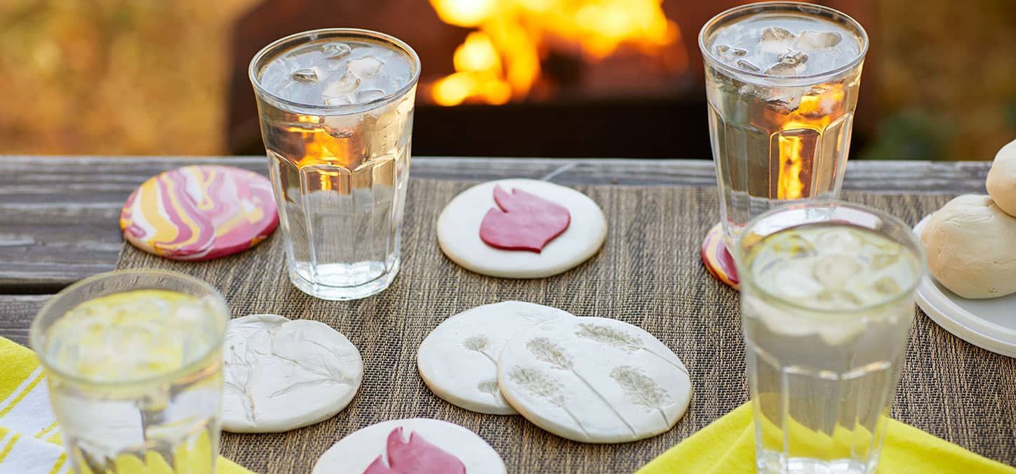 A table with a fire pit in the background and glasses of water and homemade clay coasters on it.