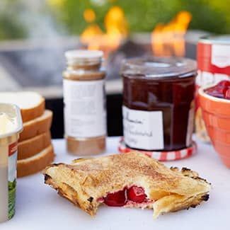 A campfire pie sandwich with a bite taken out of it is sitting on a table next to a jar of jam in front of a fire pit