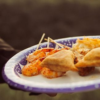 A paper plate holds shrimp skewers and fried pastries; held outdoors.