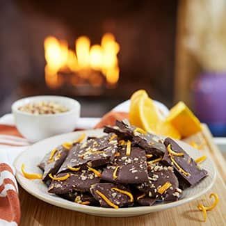 Chocolate bark with orange zest and nuts on a plate; a fire in the background.