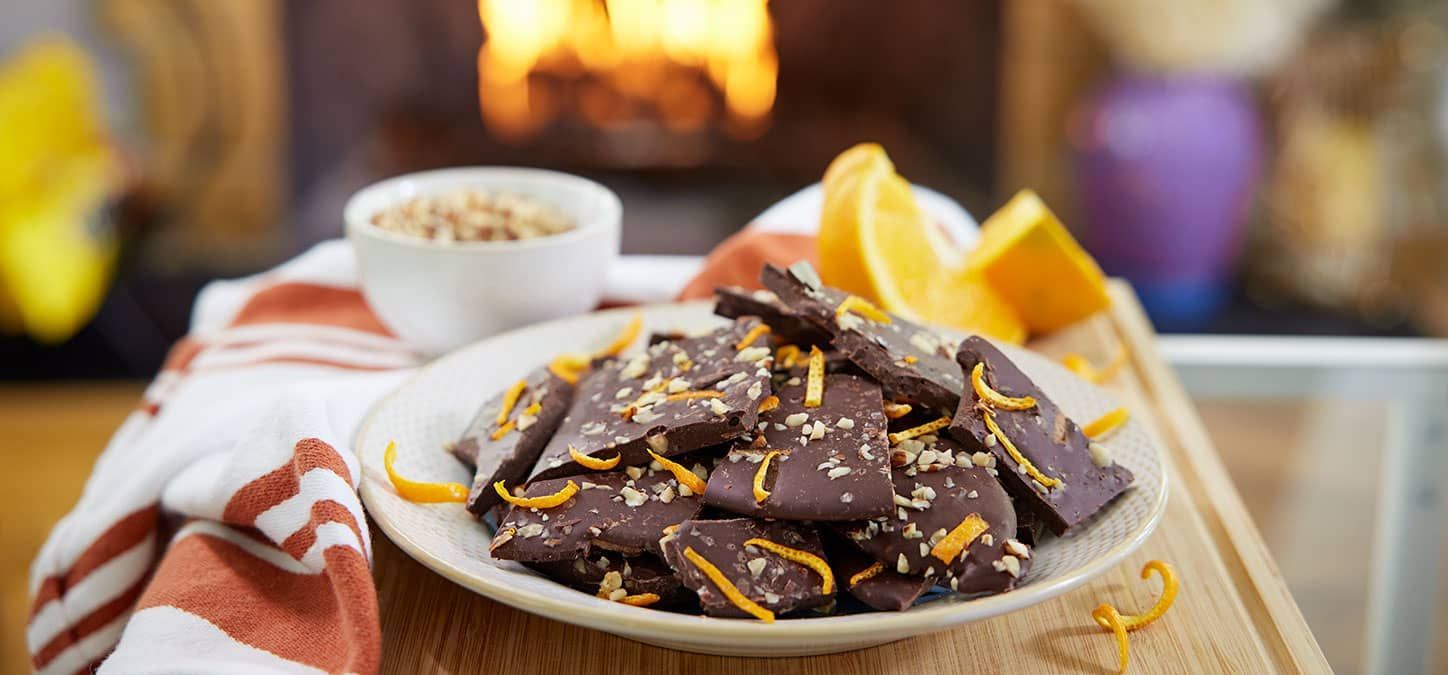 A white plate topped with chocolate bark with orange zest on a table with a fireplace in the background