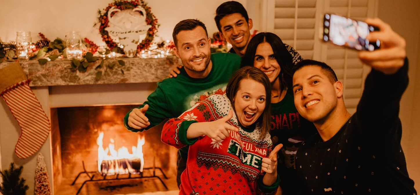 A group of people in ugly holiday sweaters are taking a selfie in front of a fireplace.