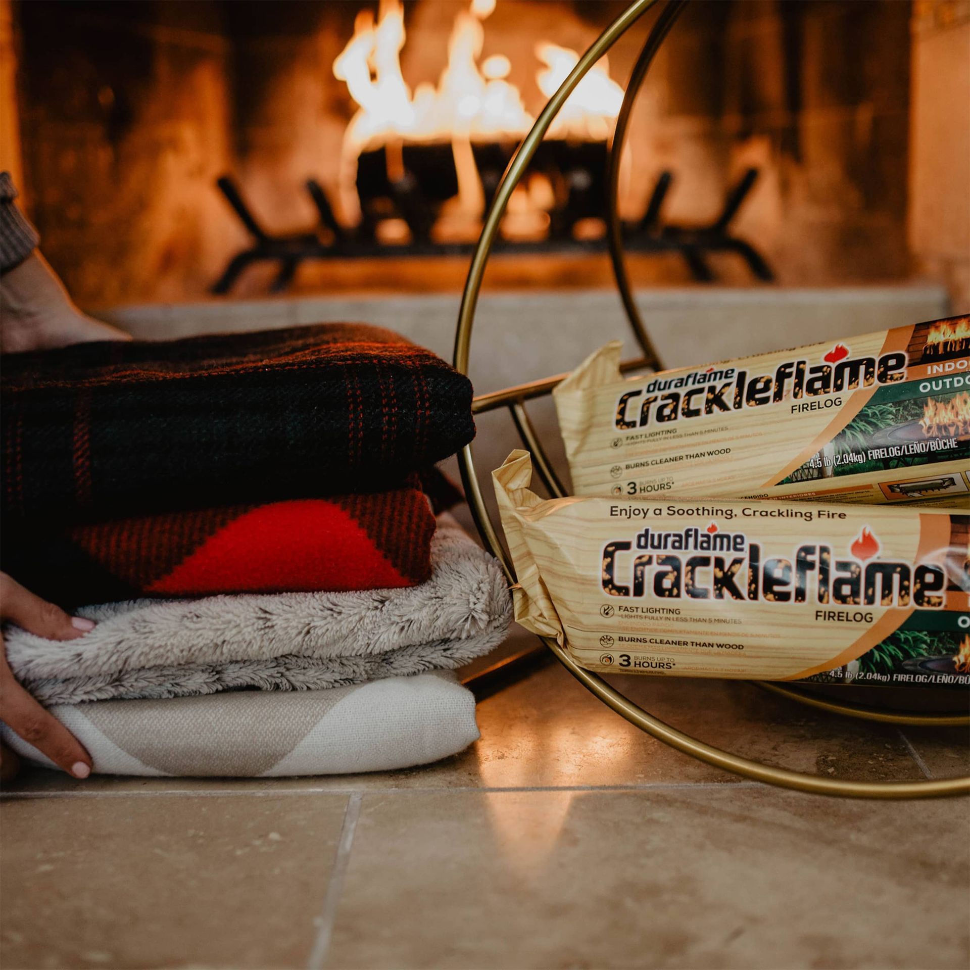 A person is holding a stack of blankets in front of a fireplace with stacked crackleflame firelogs