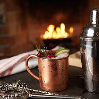 Copper mug cocktail with cranberries and lime, near a fire and cocktail shaker.