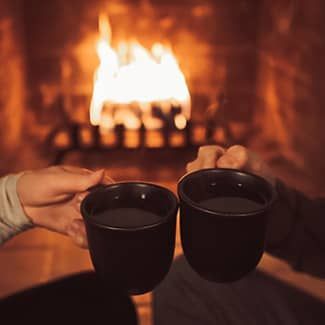 Two people are toasting with cups of tea in front of a fireplace.