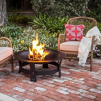 Fire pit burning brightly, flanked by wicker chairs with pillow and a blanket on a brick patio.