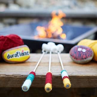 Three marshmallow sticks are sitting on a wooden table in front of a fire pit.