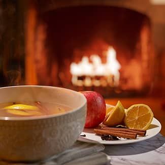 Bowl of warm drink with fruit, spices, and a lit fireplace in the background.