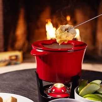 A person is dipping a piece of bread into cheese in a red pot of fondue in front of a fireplace with a duraflame crackleflame fire log in background